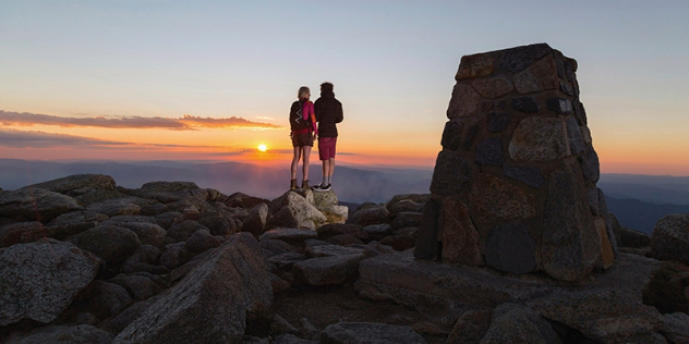 A couple stands together on a pile of rocks at the top of a mountain watching a sunset.