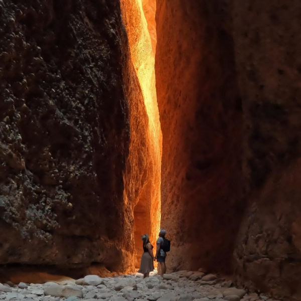 A couple holds hands at the bottom of chasm extending dozens of metres above, lit with orange sunlight.