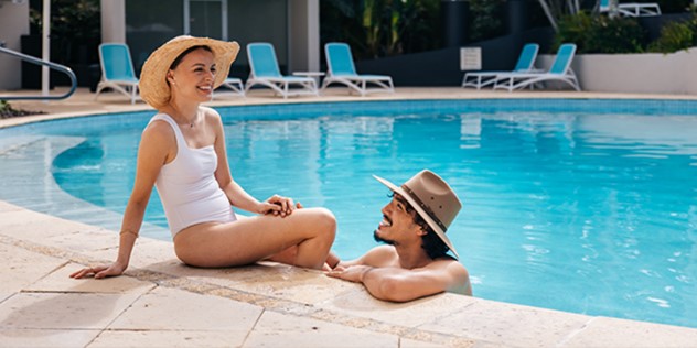 A woman in a white one-piece bathing suit perches on the curved side of a pool, surrounded by blue deck chairs, while a man in a sun hat bobs in the water next to her. 