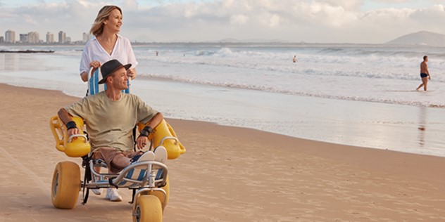 A woman pushing a man along a beach in an accessible tricycle, as waves lap at the shore, the town of Mooloolaba is visible in the distance.