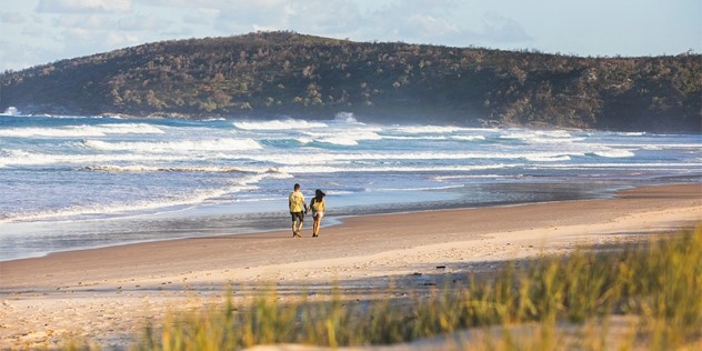 A couple in faded yellow shirts, walks down a sandy beach, with a frothy ocean of waves lapping at the shore, a green scrubby hill and cloudy sky in the background.