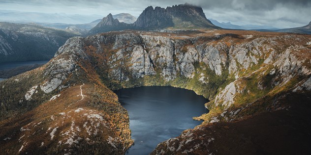 A dark blue lake at the bottom of a deep rounded pit in a mountaintop, with a mountain ranges stretching out in the distance.