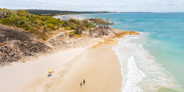 A rocky, tree covered outcrop at the end of a sandy beach with a single striped umbrella, the rest of the coastline in the distance.