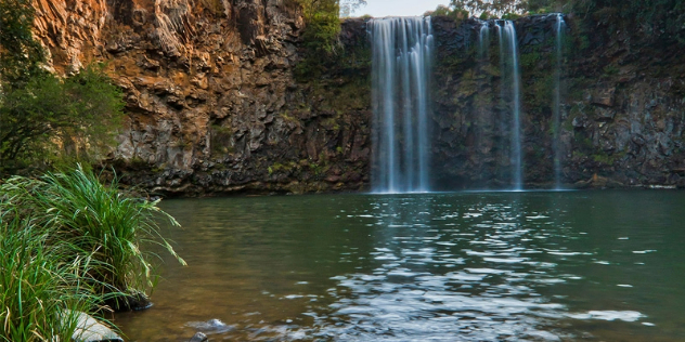 Several thin streams of water pour into a blue-green pond surrounded by a red-orange rock walls and vegetation.