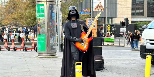 A Darth Vader cosplayer holds an electric guitar, next to a queue of people outside a convention.