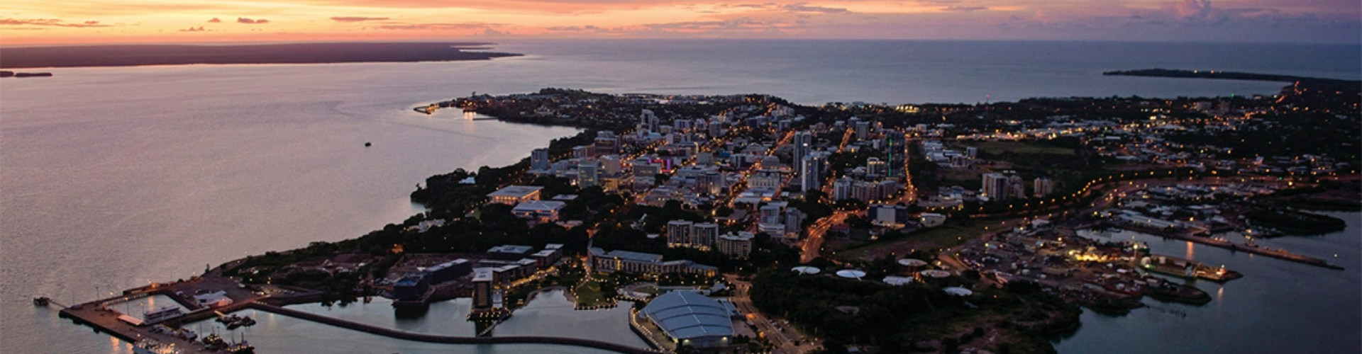 darwin-at-dusk-nt-1920x500 A near-aerial view of a city with a harbour spread along a peninsula, under a purple sunset.