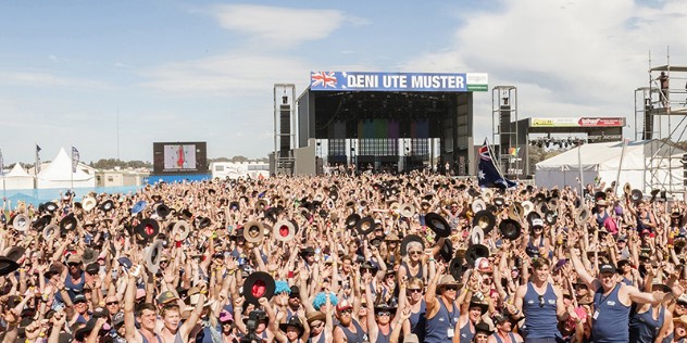 A large crowd in front of a sunny outdoor stage area, all holding a hat or their hands in the air.