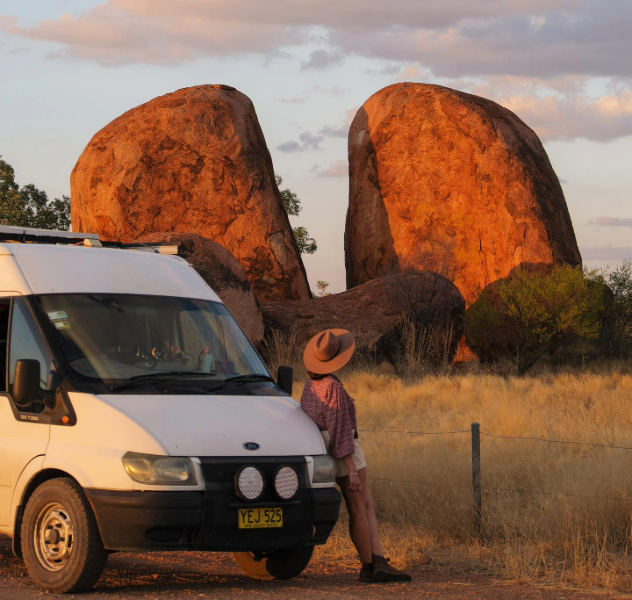 A woman leans on a van looking at two approximately ten metre tall rounded rocks lit by orange sunset.