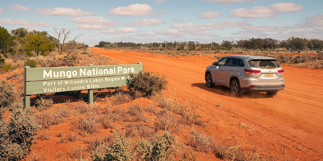  A SUV travels along a sunny, orange dirt road past a wooden sign for Mungo National Park.