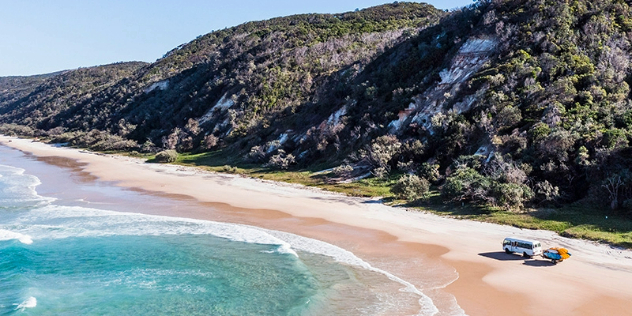 A white bus towing a trailer appears small driving along a sunny, sandy beach past tall green hills.