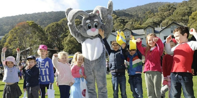 A group of kids with an Easter bunny character in a green field, with a hotel in the background.