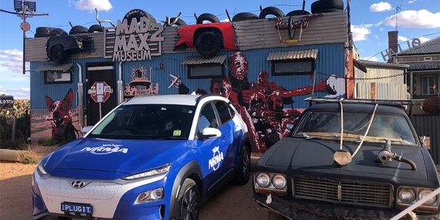 nrma electric vehicle parked next to vehicle from the mad max 2 movie outside a tin building painted with movie characters