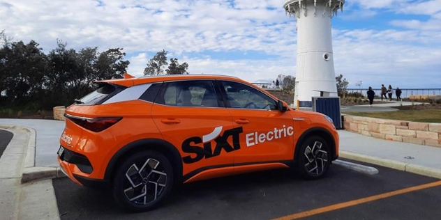 orange rental car branded SiXT Electric in parking spot with Warden Head lighthouse and coastal walk in the background