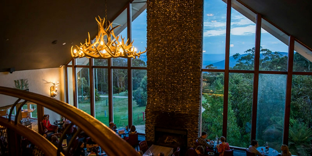  View over a balcony at a chandelier made of antlers, down into a lodge dining room with floor to ceiling windows and fireplace.