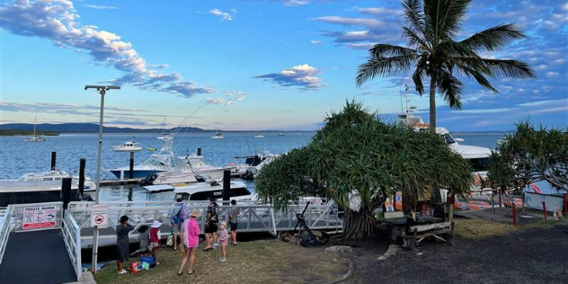  A family in hats and shorts gathers near trees and a walkway to a pier by a busy, newly-built harbour. 