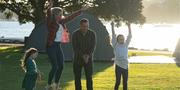 A couple and two girls jump and play on grass in the sun, near a tent set up on the water's edge.