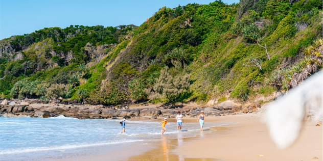 A couple and two kids walk through waves along a sandy beach leading to tall green covered cliffs and blue sky.