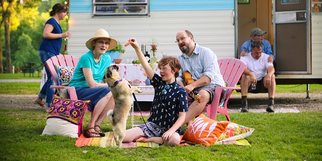 A child and his family play with a dog on the grass in front of a holiday trailer campsite. 