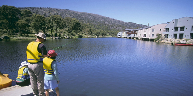 A man and child in lifejackets stand fishing off a concrete pier into a lake with buildings on stilts on the opposite shore.