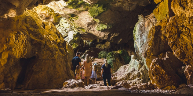 A couple and three young girls stand in a rocky, mossy cave lit by sunlight coming through above.