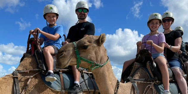  A couple and two children in helmets riding camels, framed by blue sky. 