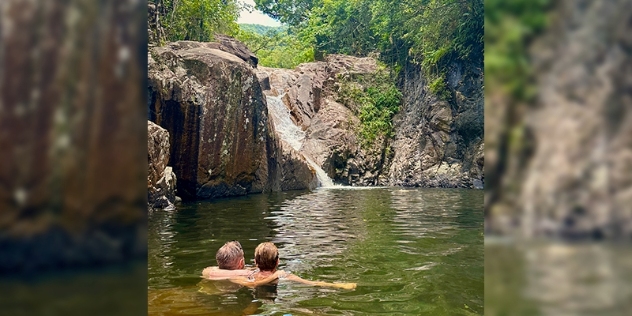 couple relax in secluded natural pool surrounded by rock and bush and fed by a small waterfall