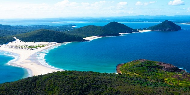 A flyover view of a deep blue tropical ocean bay created by green hills divided by a white sandbar, where the water turns turquoise as it hits the sandbar shores, under a blue sky.