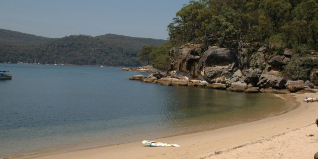 A small sandy beach, rising into a rocky, treed shoreline as it goes out into the wider bay, surrounded by green hills and blue sky.