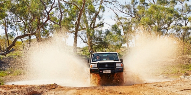 a four-wheel drive vehicle sprays water as it emerges from a pool of water in the dip of a dirt road 