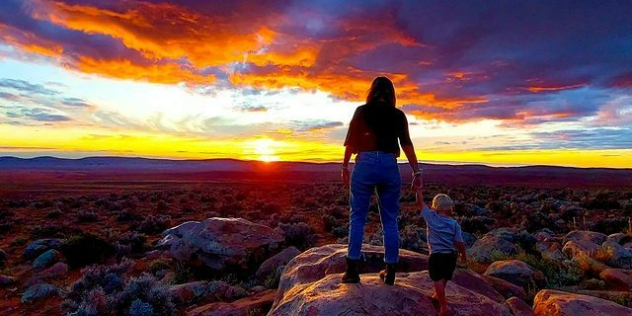 A woman and toddler on a rock watch the sunset over flat red scrubby earth and mountains on the horizon. 