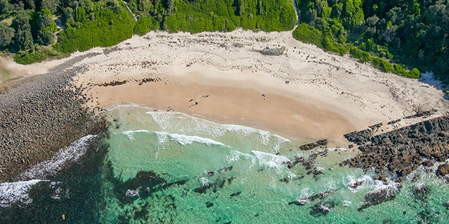 arial view of waves breaking on a section of sandy beach with rocks to the right and pebbles to the left