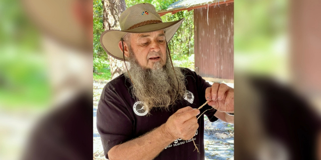 An Aboriginal man with a beard, in a brimmed hat, holds out a strand of twine.