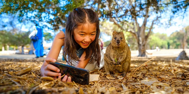  A girl gets on the ground to take a selfie with groundhog-like marsupial whose expression looks like a smile.