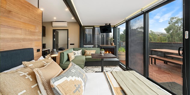 A bed in the foreground, with a L-shaped couch, small table, kitchen and fireplace, all facing the opposite wall covered with glass sliding doors.