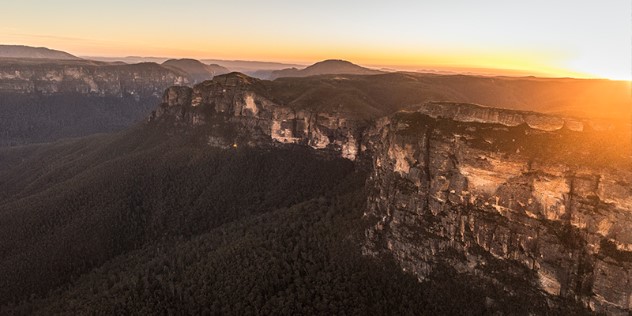 A warm orange sunrise breaks over a vista of the rocky cliffs that lead into the tree-lined valley of Govetts Gorge. 