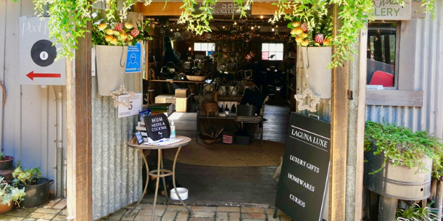  Sunlight hits vines hanging over open corrugated iron doors leading into a warm lit wood lined showroom of artisan products on display.