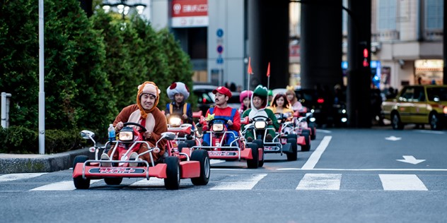 A group of six people in Mario video game character costumes, driving go carts on a sunny day.