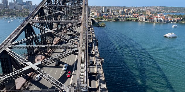 A close-up view of the Sydney Harbour Bridge gridwork and cars passing below from the perspective of the Pylon Museum lookout. The opposite shoreline is lined with residential homes and boats passing through the harbour. 