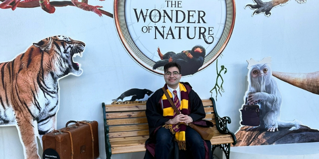 A smiling man in a Harry Potter costume, sitting on a bench outside an exhibition.