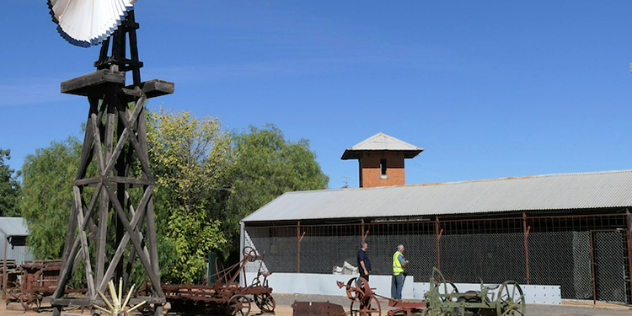  Two older men stand among old farm machinery, a windmill, and long building with wire mesh along the front.