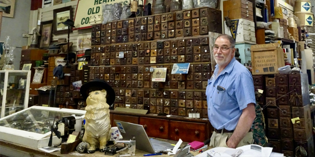  An bearded man stands behind a counter and in front of a large wall of tiny drawers and antiques.