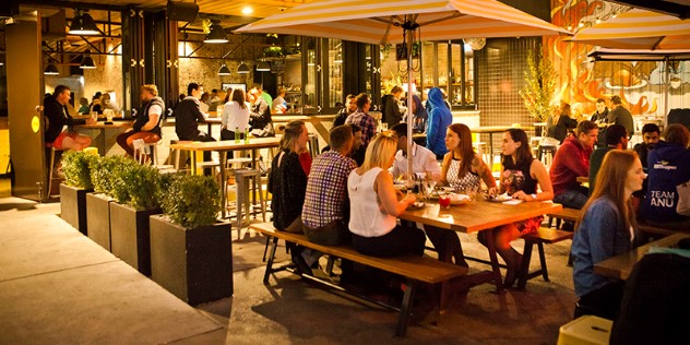 A photo of the patio of Hopscotch bar in Canberra, groups of people sit at tables under umbrellas and at the bar, under the warm glow of overhead lamps.