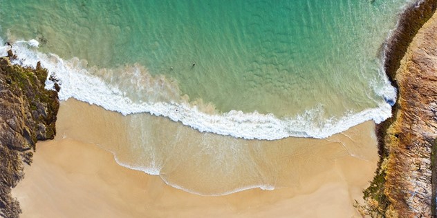 An aerial view of a sandy beach on a sunny day, framed by two rock walls, as a green blue ocean wave washes on the shore, dividing the frame with white froth. 