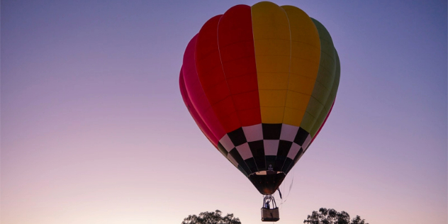 A single hot air balloon with a vertical rainbow pattern on top and a black and white checkered section just above the basket, floating above treetops, against a purple sky.