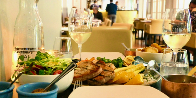  A table with a plate of fish and two glasses of white wine, in a warmly lit, historic dining room with tall windows on one side.