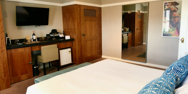  A hotel room with a bed and warm wood cupboards along one wall with an inset desk and historical items like a hat on top of it.