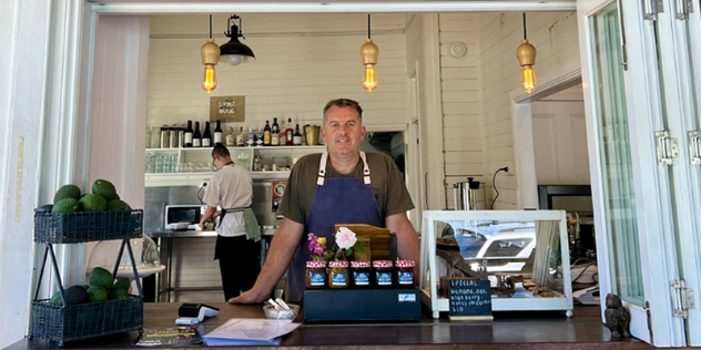  A short-haired man in an apron leans out of a white, wood cafe window behind a display of baked goods and jams.