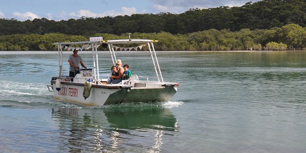 A woman and two kids ride a small, jetboat style ferry along an inlet. Thick foliage lines the shoreline.