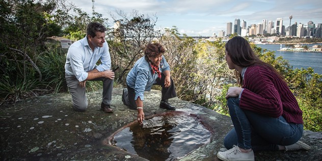 A man and a woman sit listening to an indigenous woman speak as she crouches and points to a water-filled rounded indent in rocks along the shore opposite the sunny Sydney harbour and city skyline. 