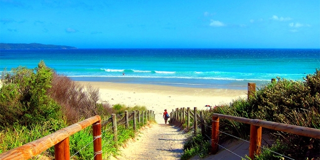 a surfer wearing a red rashy and carrying a blue surfboard walks along a sandy boardwalk flanked by low shrubs and grasses that slopes down to a white sandy beach and turquoise ocean